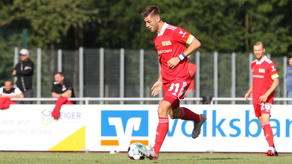 A soccer player in a red jersey is dribbling the ball on a practice field. Spectators in the background.