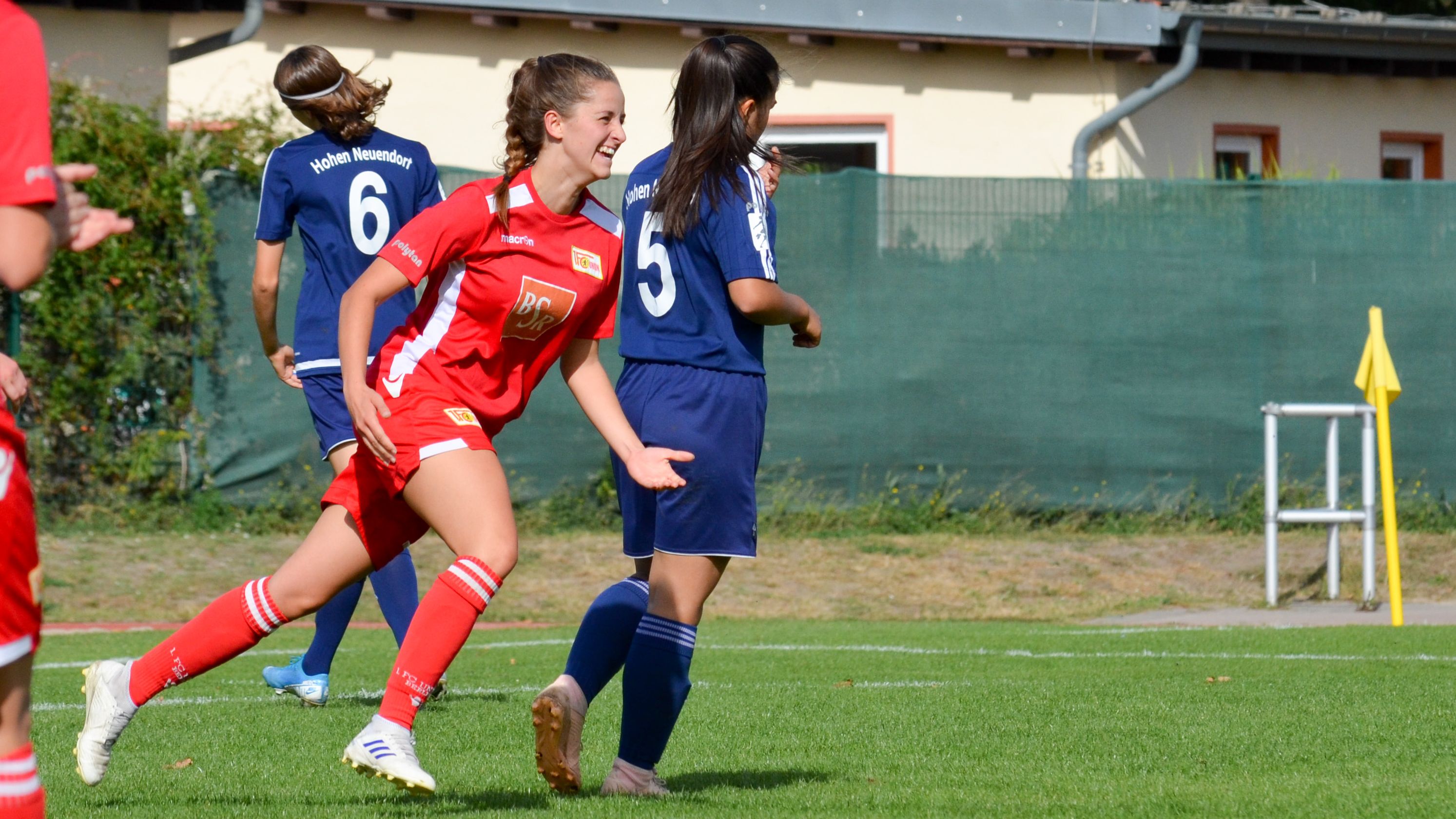 Ein Fußballspiel, bei dem eine Spielerin in rotem Trikot jubelt, während andere Spielerinnen in blauen Trikots auf dem Feld sind.