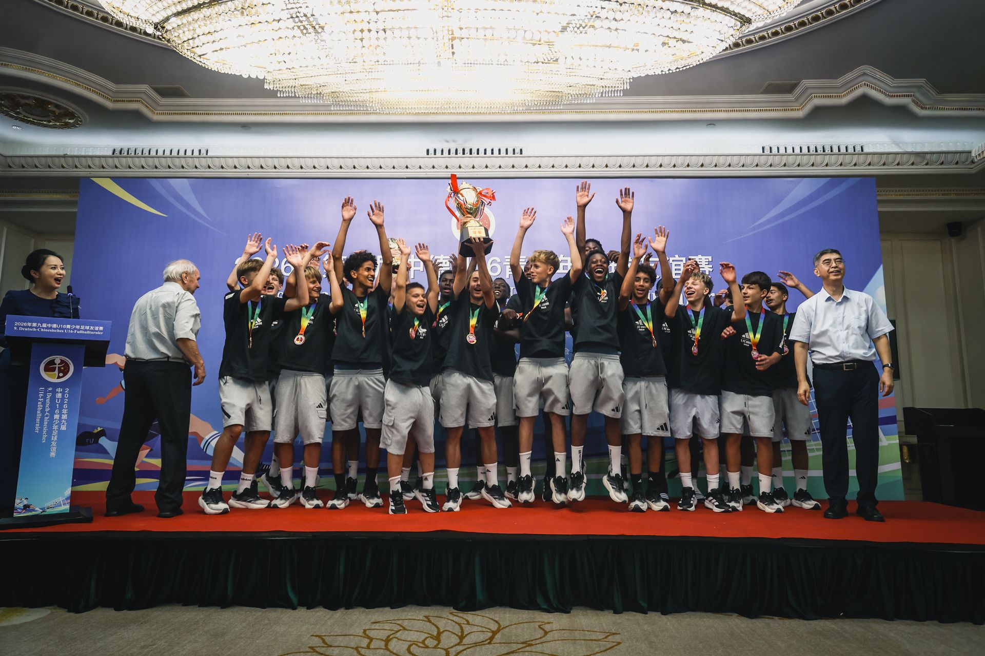 Gruppe von jungen Sportlern in schwarzen T-Shirts und Shorts jubelt auf einer Bühne mit einem Pokal und Medaillen.