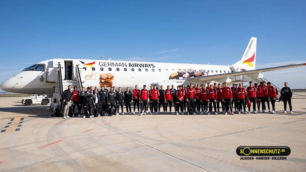 Eine Gruppe von Sportlern steht vor einem Flugzeug mit dem Schriftzug "GERMAN AIRWAYS" auf einem Rollfeld.