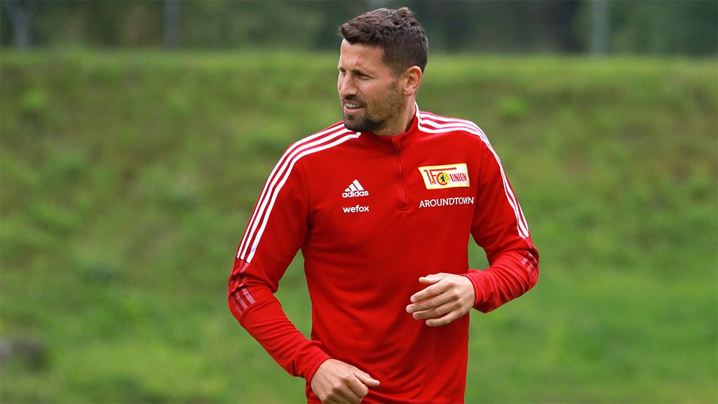 A football player in a red tracksuit with white stripes and the club logo on a sports field.