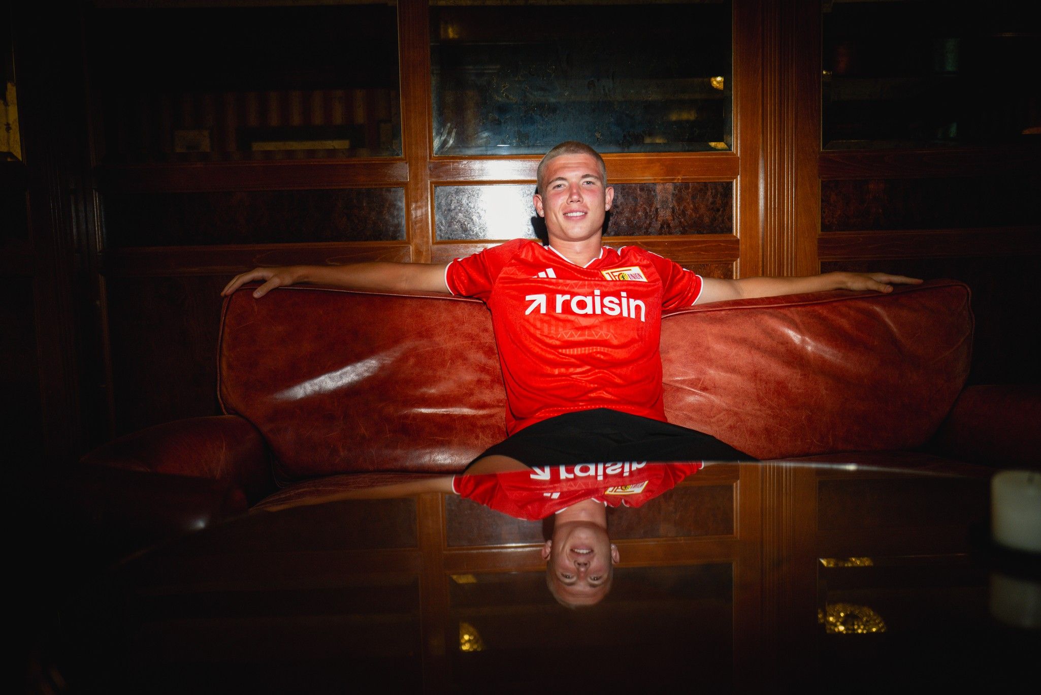 Ein lächelnder junger Mann in einem roten Fußballtrikot sitzt entspannt auf einer Couch, der Tisch spiegelt sein Bild wider.