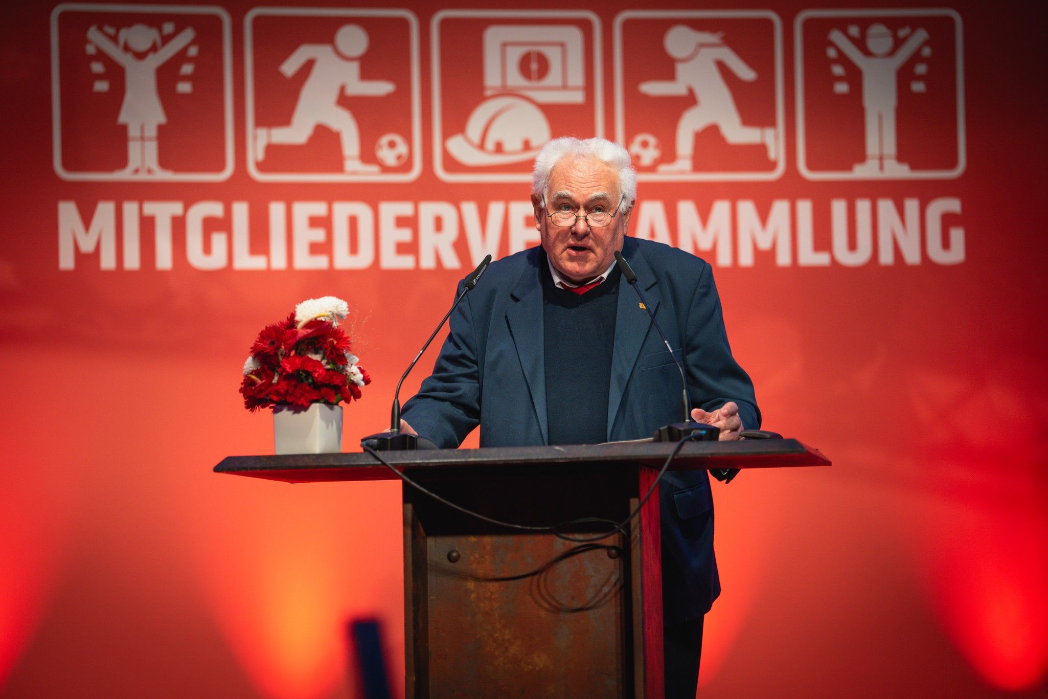 Älterer Mann spricht an einem Rednerpult während einer Versammlung, Hintergrund mit grafischen Symbolen und roter Beleuchtung.