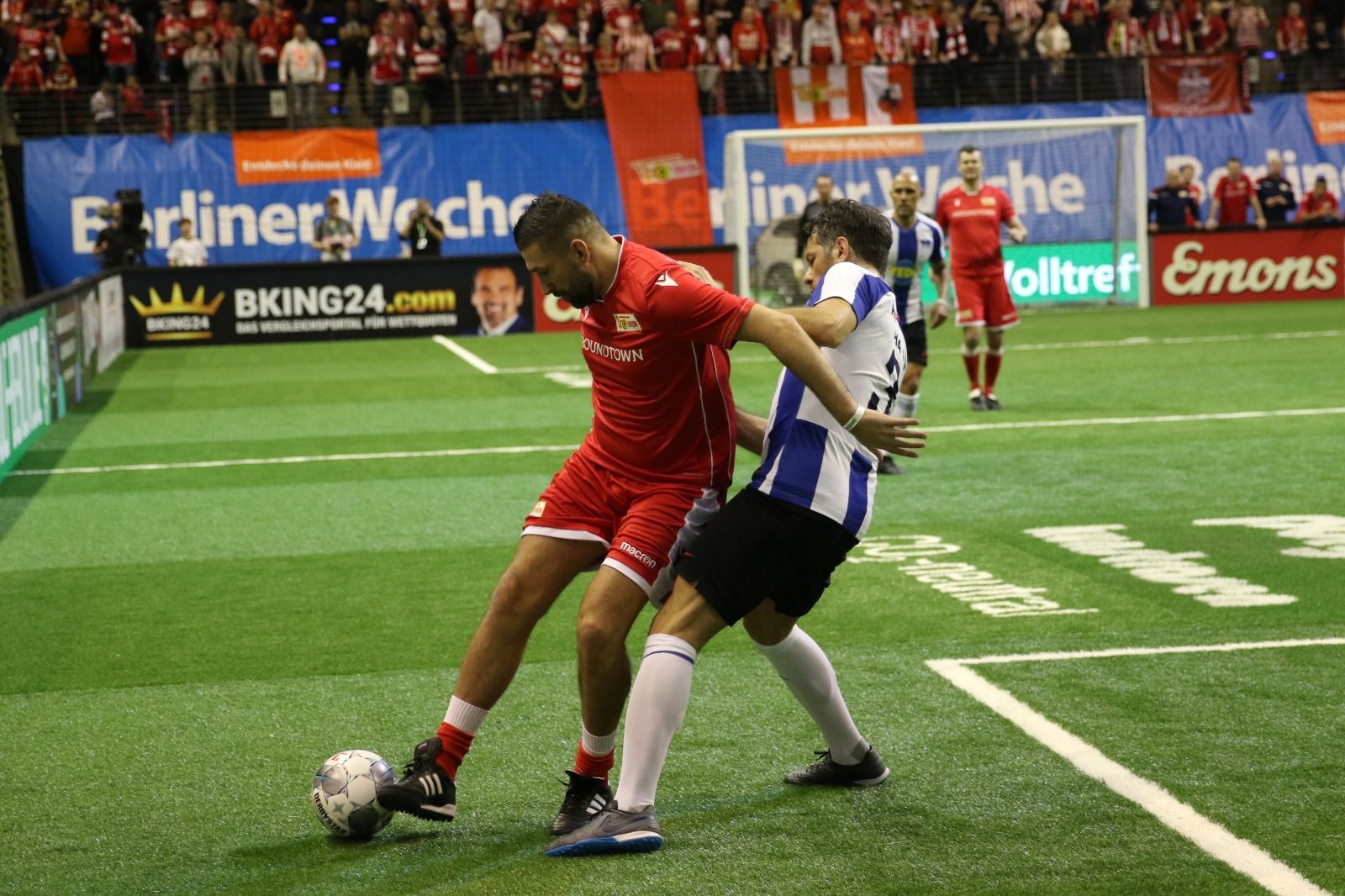 Two soccer players are battling for the ball during a game in a hall. Spectators are visible in the background.