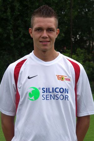 Junger Fußballspieler in einem weißen Trikot mit einem Logo und Sponsorenaufdruck, steht auf einem Sportplatz.