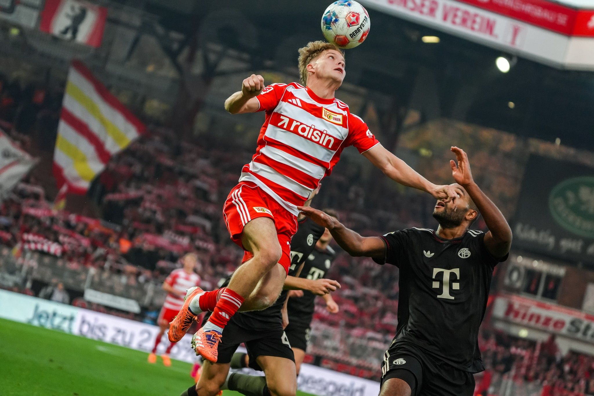 Spielszene im Fußball mit einem Spieler, der hochspringt, um den Ball zu köpfen, während ein Gegner ihn anvisiert.