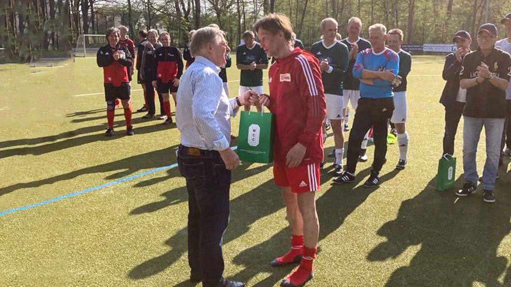 Ein Fußballspieler in rotem Trikot erhält einen Preis von einem Mann in Hemd, umgeben von Zuschauern auf dem Platz.