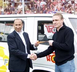 Zwei Männer schütteln sich die Hände vor einem Auto, während eine Menschenmenge im Hintergrund steht.