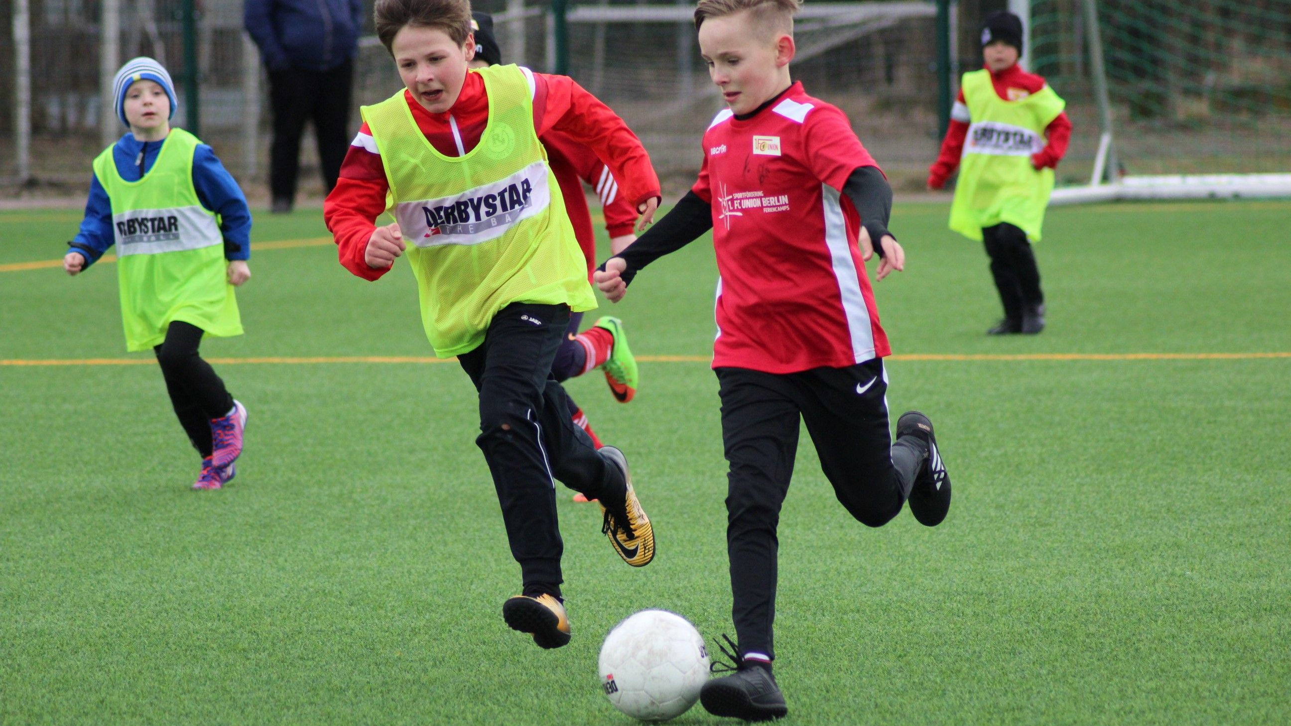 Zwei Kinder in roten und grünen Trikots spielen Fußball auf einem Kunstrasenplatz.