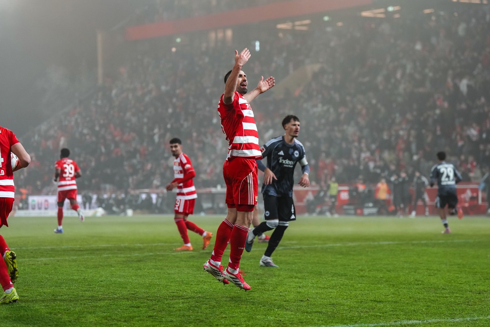 Fußballspieler in roten und weißen Trikots jubeln auf dem Spielfeld vor einer jubelnden Menge im Hintergrund.