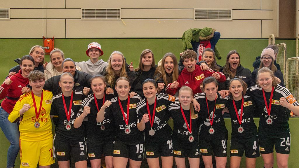 Gruppenszene von Fußballspielerinnen und Trainerinnen in Schwarz mit Medaillen, lächelnd und mit erhobenen Fäusten, in einer Sporthalle.