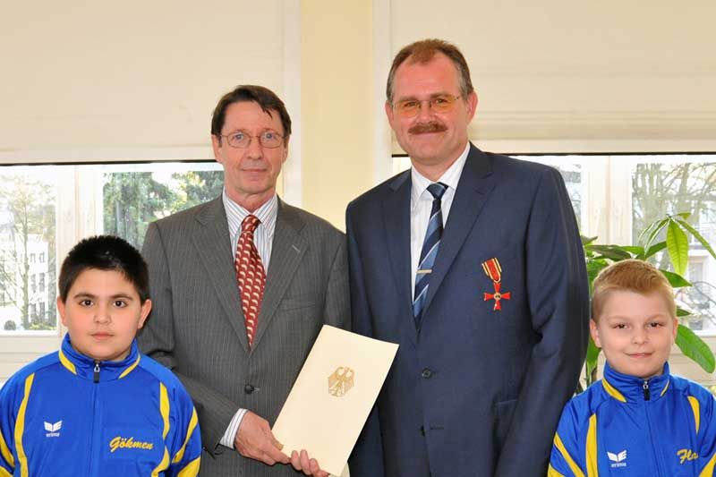 Vier Personen in einem Raum, darunter zwei Kinder in sportlicher Kleidung und zwei Erwachsene in Anzügen, mit einer Urkunde in der Mitte.