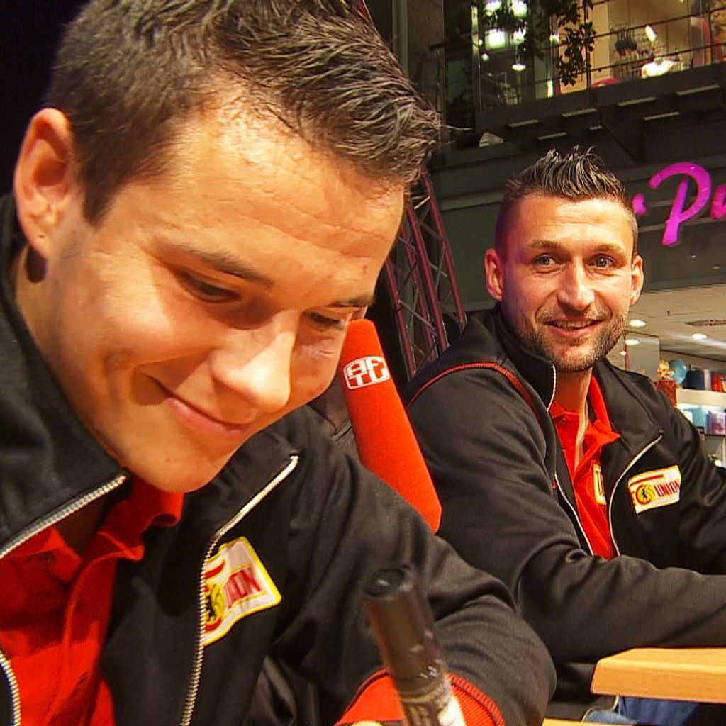 Zwei männliche Personen in rot-schwarzen Oberteilen sitzen an einem Tisch und lächeln, während sie etwas auf einem Blatt Papier schreiben.