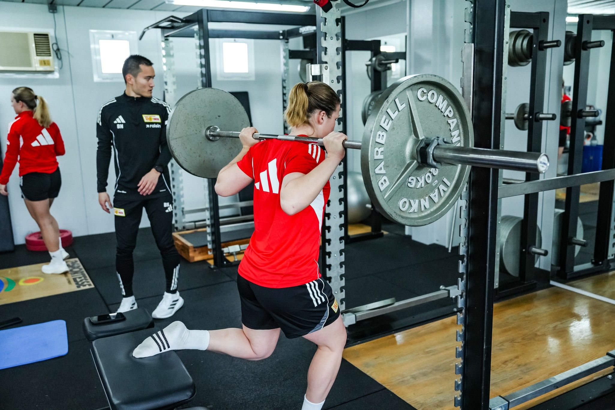 Eine Frau in rotem Shirt macht eine Kniebeuge mit einer Langhantel, während ein Trainer zusieht.
