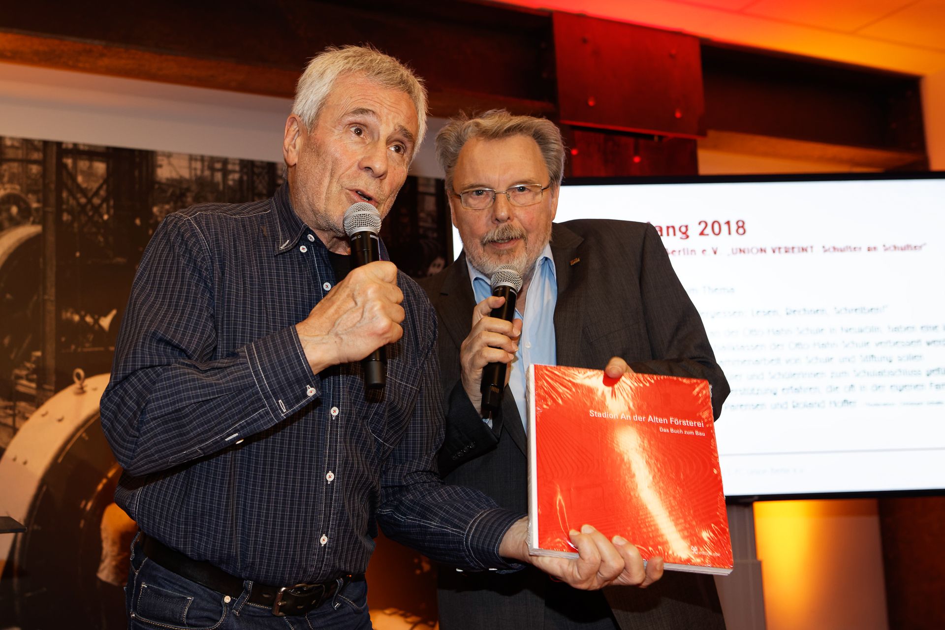 Zwei Männer stehen nebeneinander und halten ein rotes Buch. Sie sprechen in ein Mikrofon auf einer Bühne.