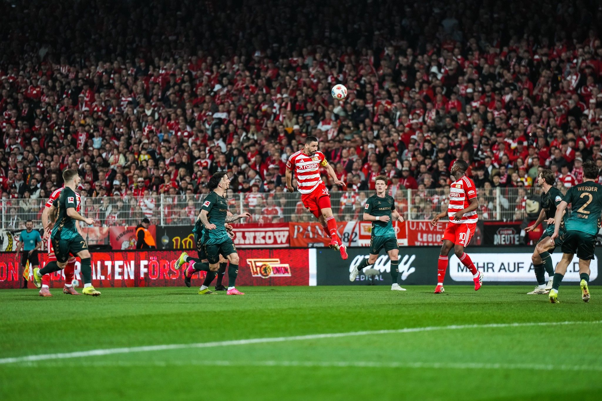Ein Fußballspieler springt hoch, um einen Ball während eines Spiels zwischen zwei Mannschaften zu köpfen, umgeben von jubelnden Fans.