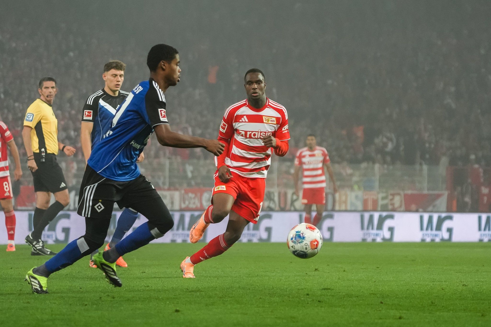 Zwei Fußballspieler im Wettkampf um den Ball während eines Spiels in einem Stadion mit jubelnden Fans im Hintergrund.
