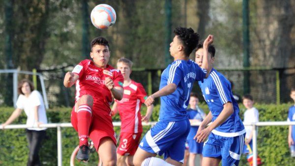 Ein junger Fußballspieler in rotem Trikot hebt den Ball mit dem Fuß an, während zwei Spieler in blauen Trikots ihn herausfordern.