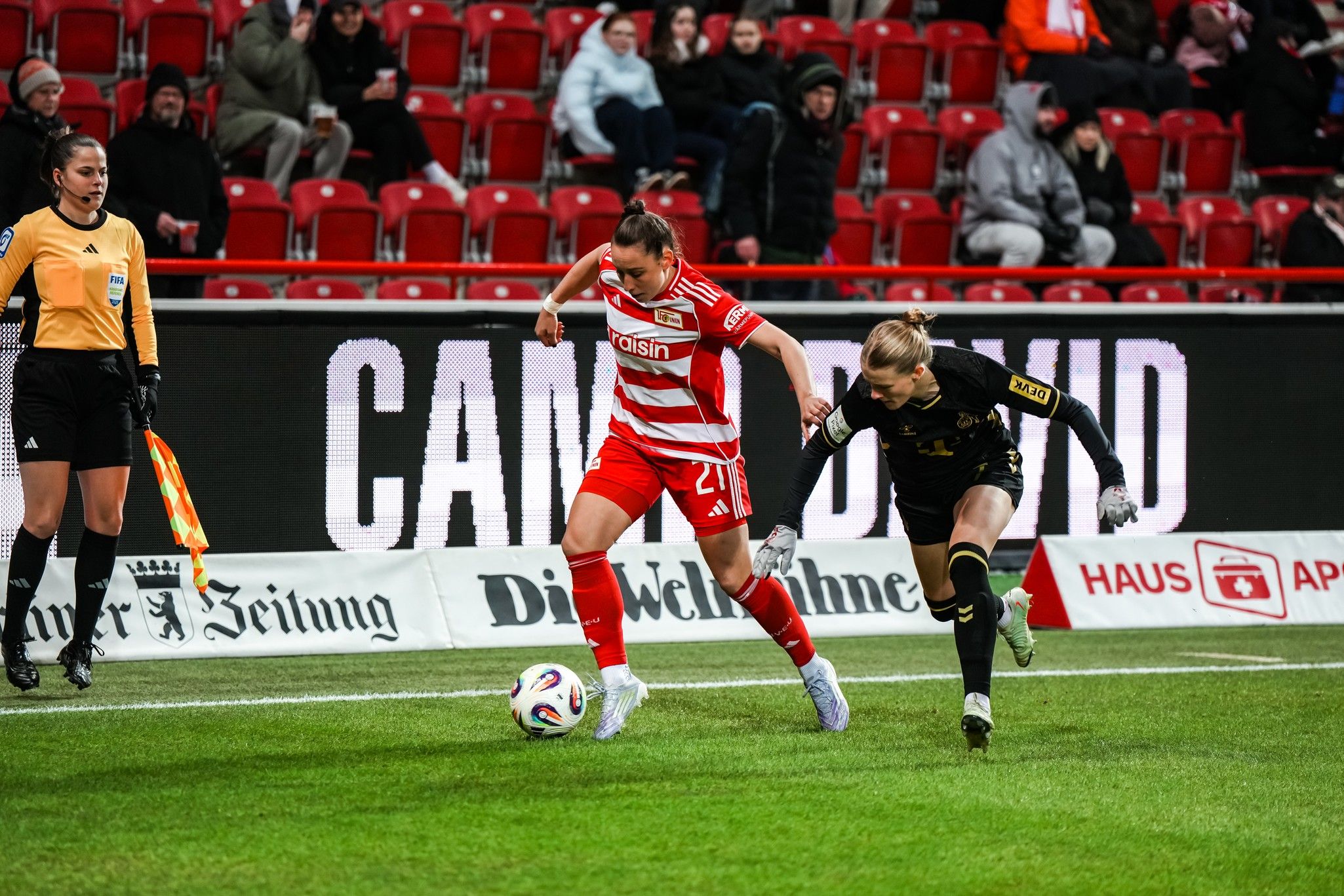 Eine Fußballspielerin in roter Uniform dribbelt den Ball an einer Torhüterin in schwarzer Uniform vorbei auf einem Stadionfeld.