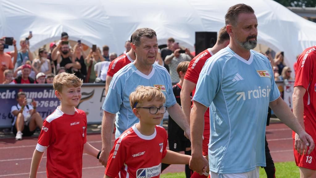 Zwei Männer und zwei Kinder in Fußballtrikots gehen gemeinsam auf das Spielfeld, während Zuschauer im Hintergrund jubeln.