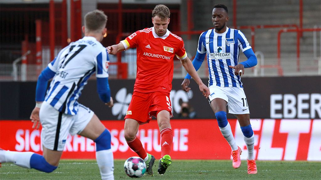 A soccer player in a red uniform is dribbling the ball while two players in blue and white jerseys try to defend against him.