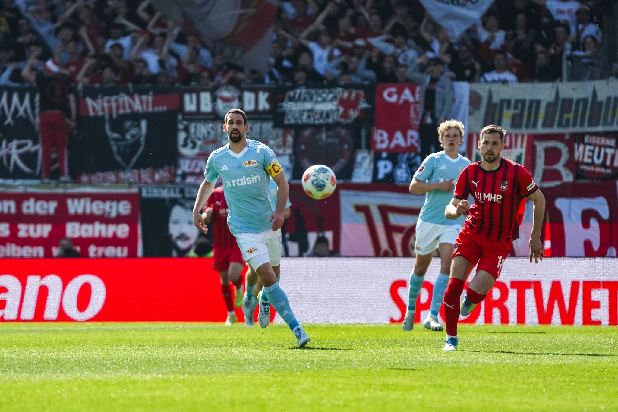 Ein Fußballspiel mit Spielern in hellblauen und roten Trikots, im Hintergrund sieht man Fans und Banner.