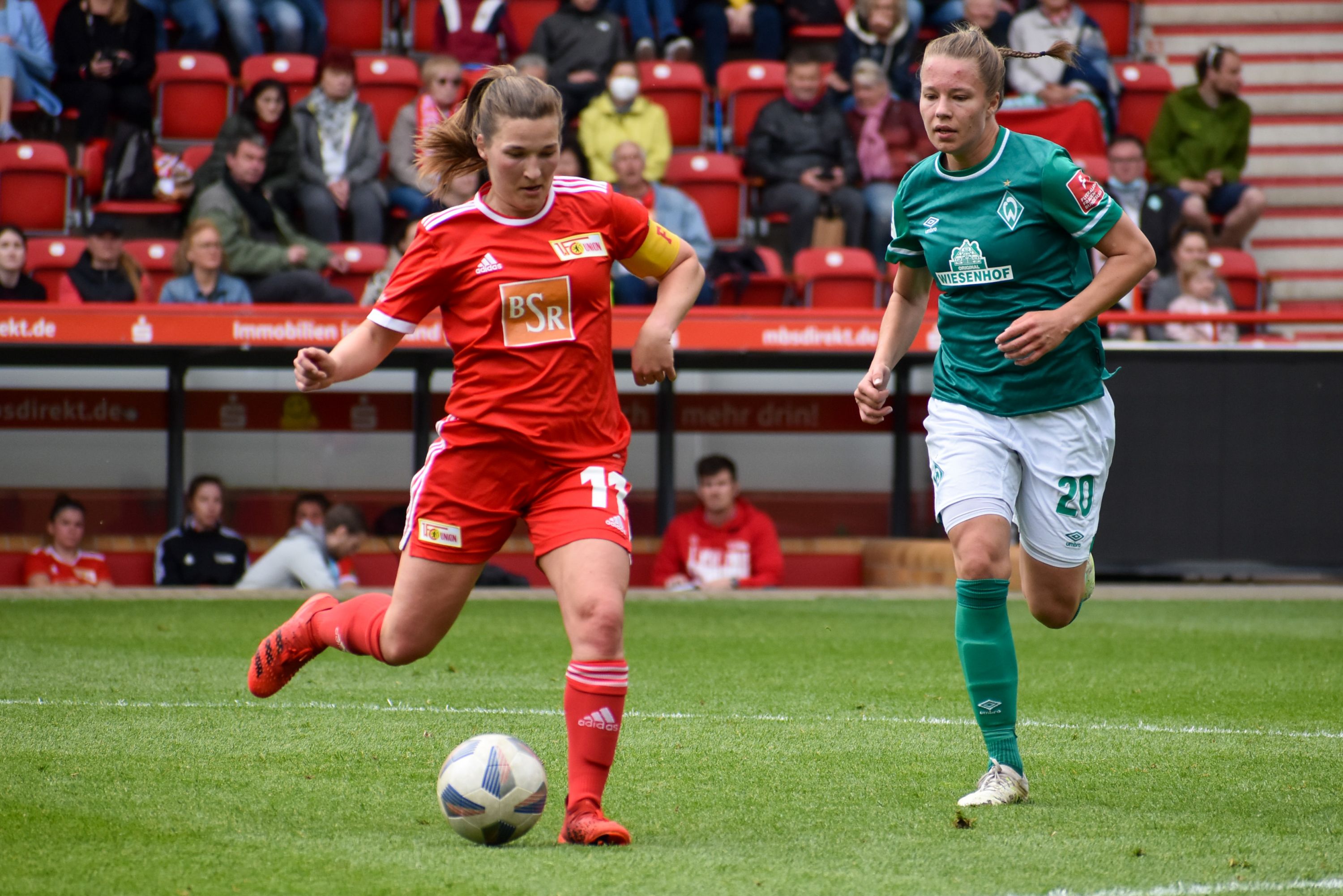 Zwei Fußballspielerinnen in roten und grünen Trikots, die in einem Stadion einem Ball hinterherlaufen. Zuschauer im Hintergrund.