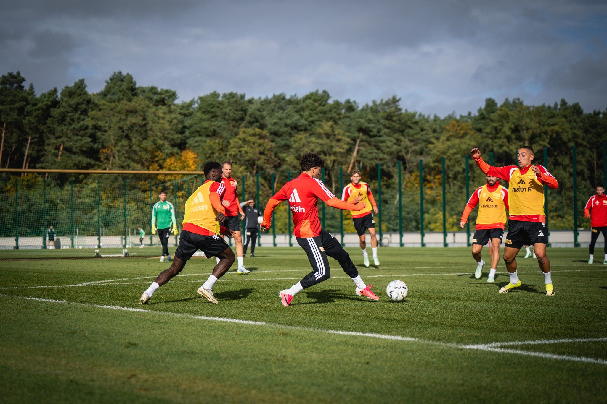 Fußballtraining mit Spielern in roten und gelben Trikots auf einem grünen Platz, umgeben von Bäumen.