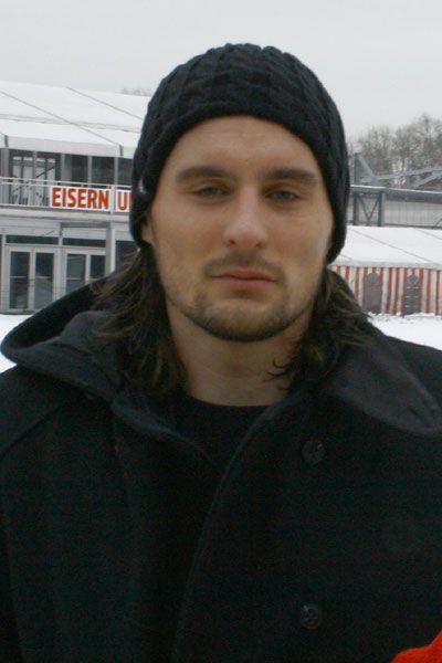 Ein Mann mit langen, dunklen Haaren und einer Mütze im Freien, vor einem Gebäudekomplex mit Schnee im Hintergrund.