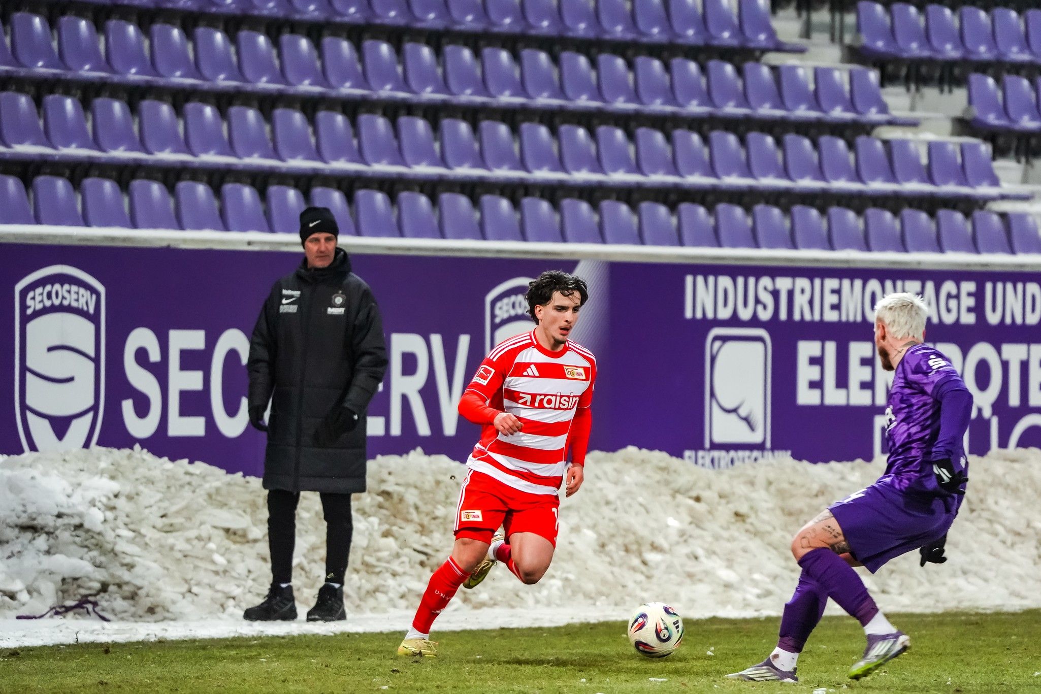 Fußballspiel: Ein Spieler mit roten und weißen Streifen dribbelt in Richtung des Torwarts, im Hintergrund schneebedecktes Feld.