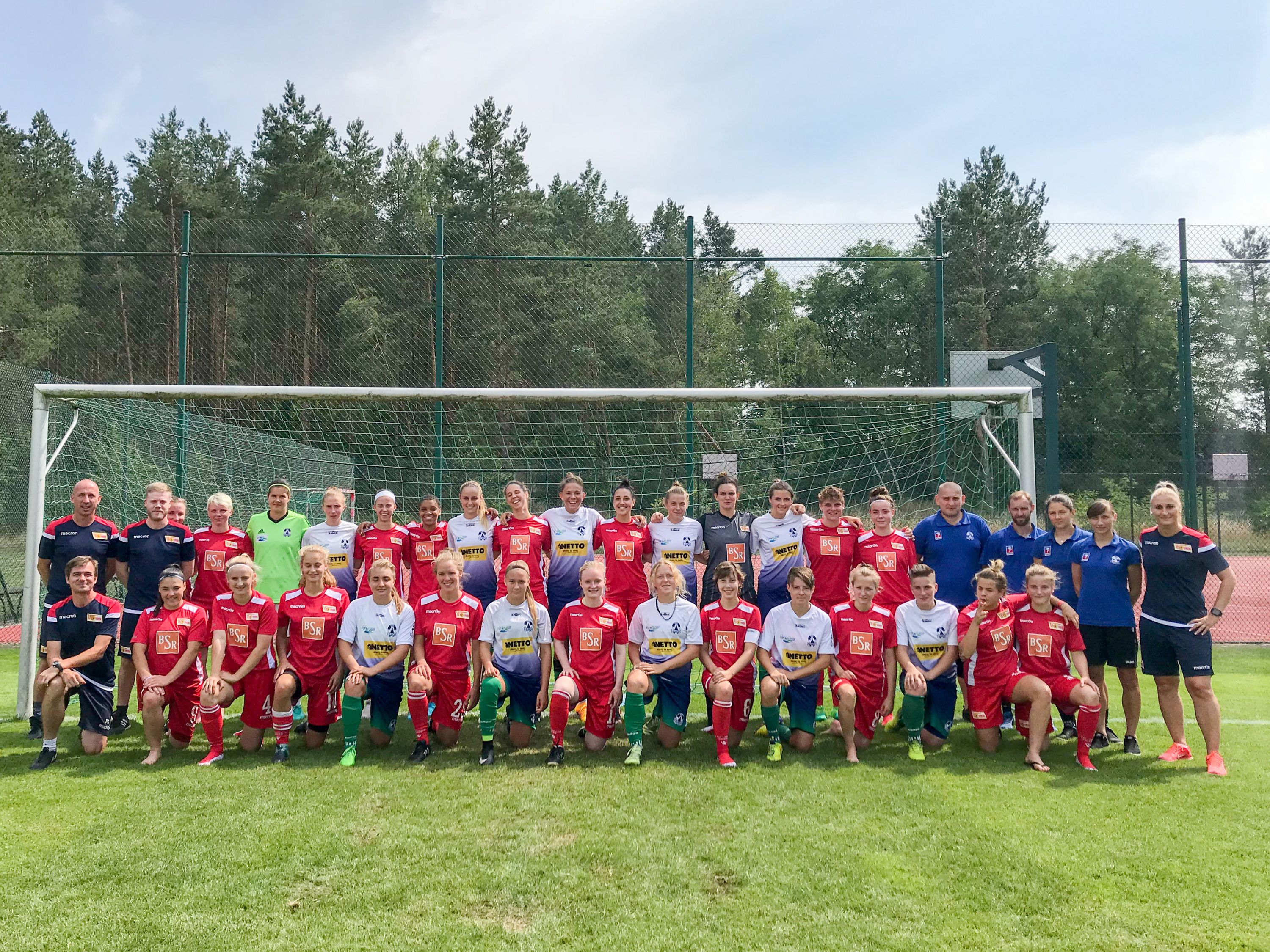 Gruppenfoto einer Fußballmannschaft mit Coaches auf einem Sportplatz, umgeben von Bäumen und einem Tor im Hintergrund.