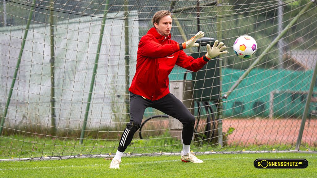 A soccer goalkeeper in a red jacket catches a ball in front of a goal during training on a green grass field.
