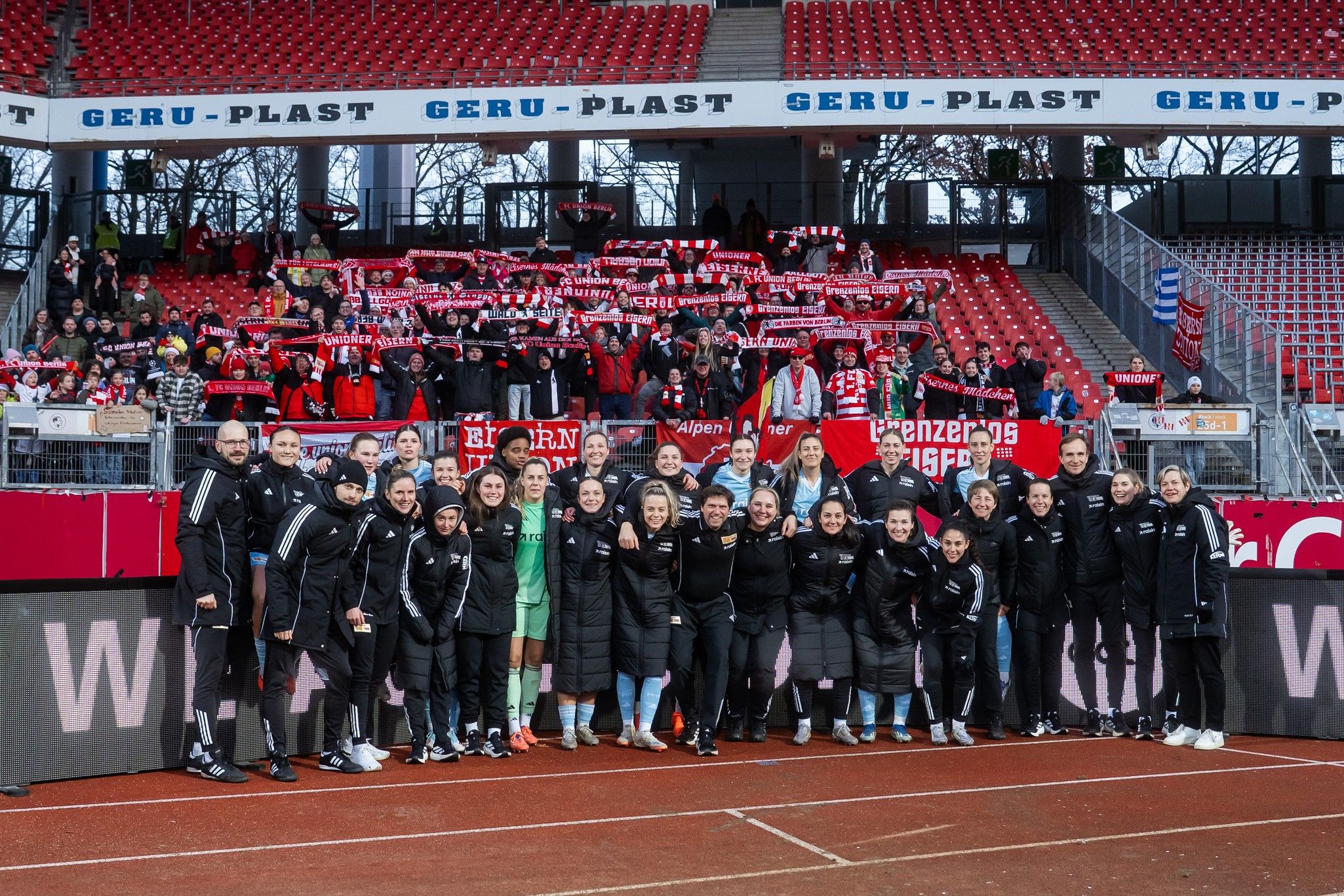 Ein Fußballteam steht lächelnd auf dem Platz, im Hintergrund Fans mit Schals in der Hand in einem leeren Stadion.
