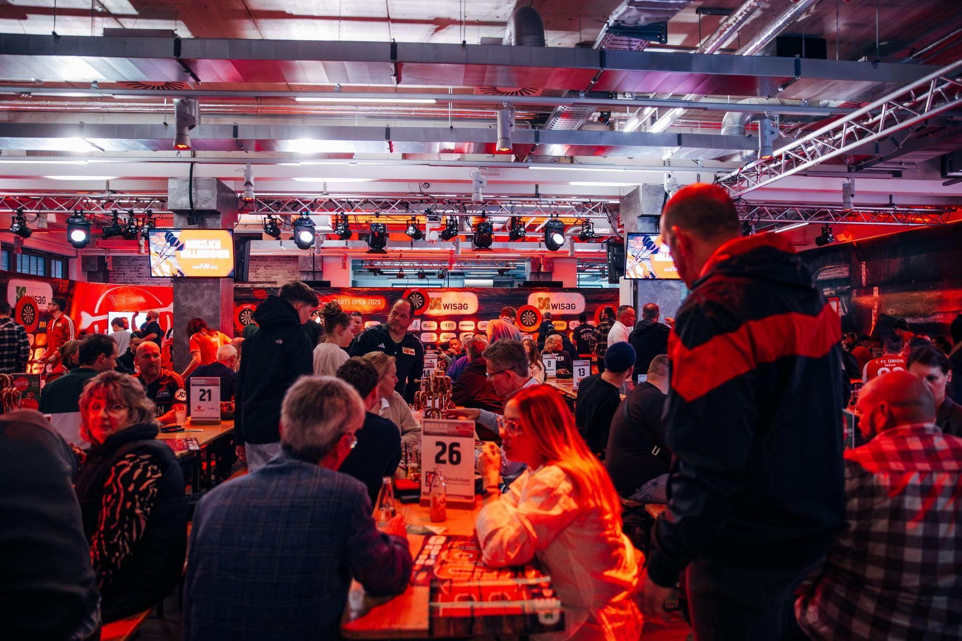 Belebte Veranstaltungshalle mit Menschen in geselliger Runde, Tischen und Bühnenlichtern, in einem festlichen, roten Ambiente.