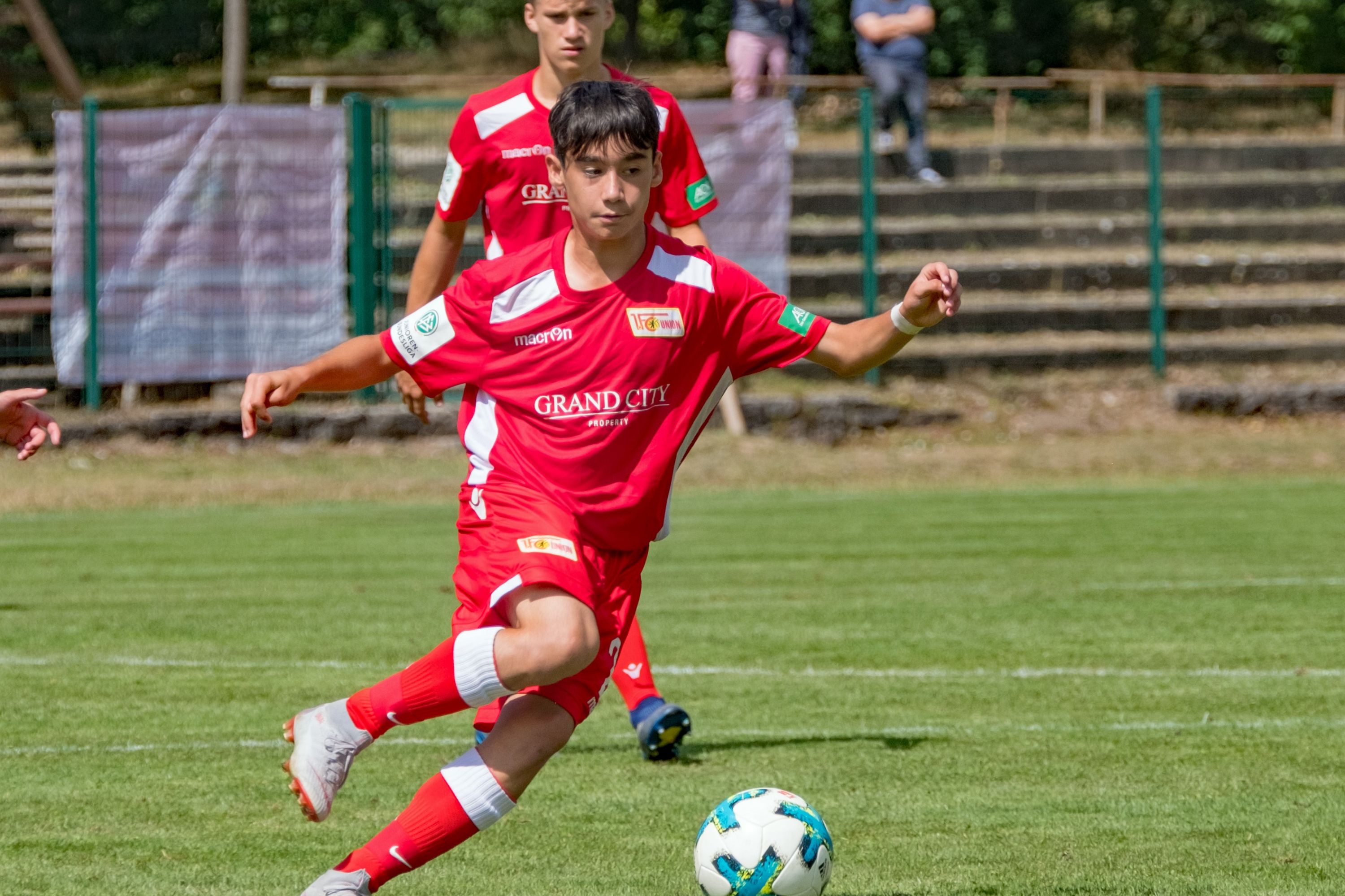Ein junger Fußballspieler in rotem Trikot tritt einen Ball auf einem Rasenplatz während eines Spiels.