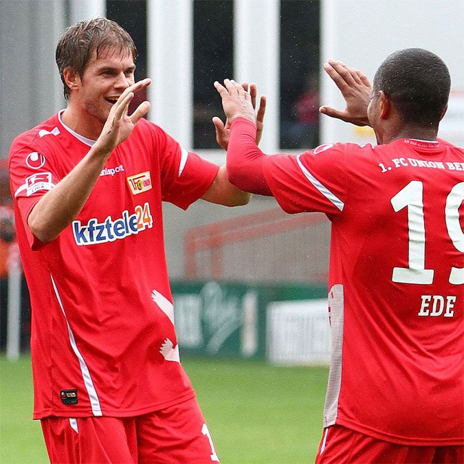 Zwei Fußballspieler in roten Trikots feiern einen Anlass, lächeln und geben sich die Hände.