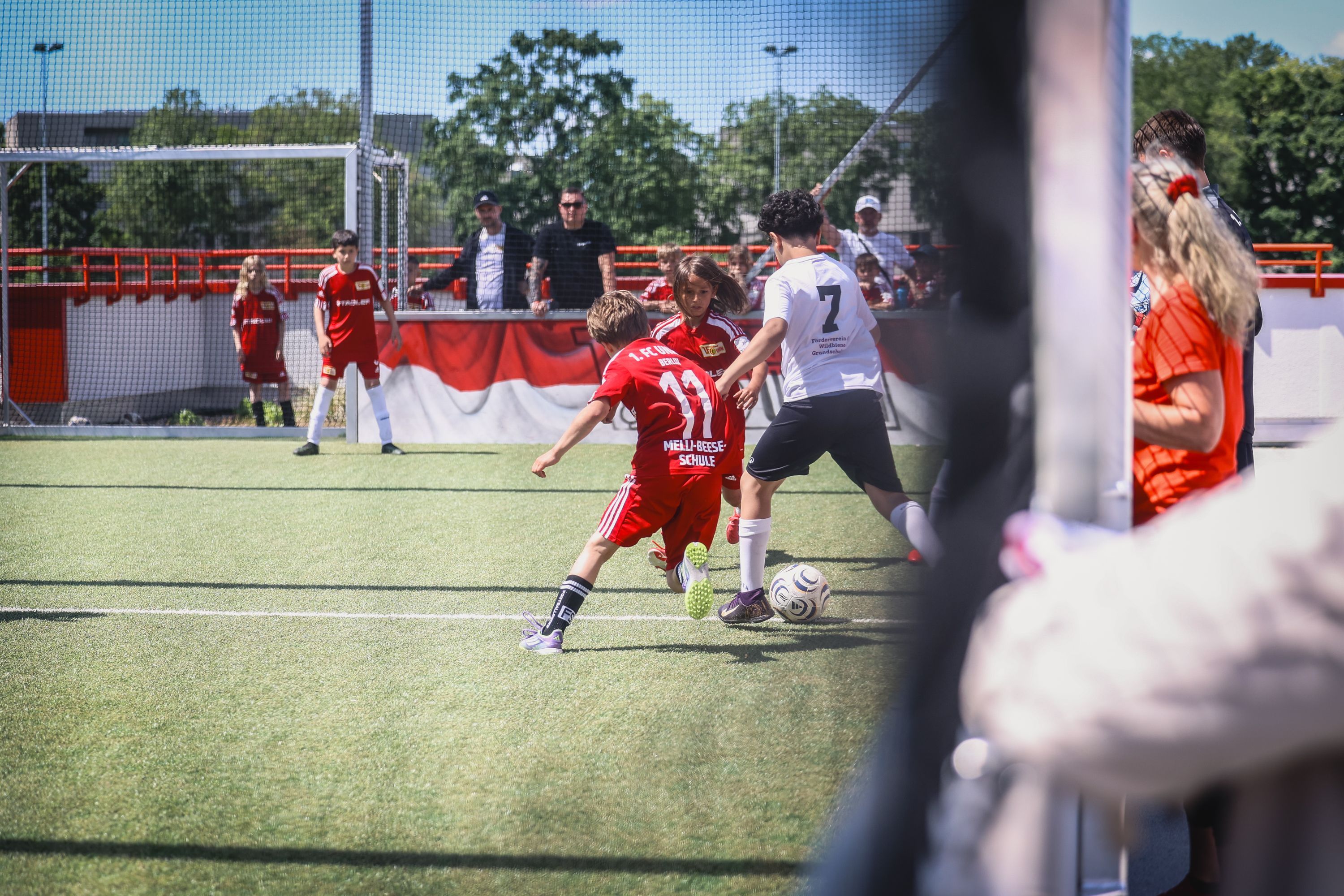 Zwei Kinder spielen Fußball auf einem Kunstrasenplatz, während Zuschauer im Hintergrund beobachten.