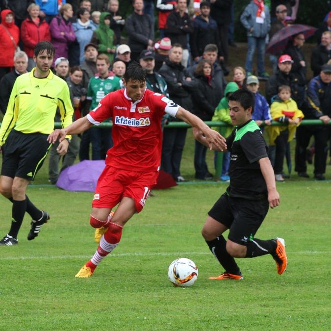 Zwei Fußballspieler kämpfen um den Ball auf einem Rasenplatz, umgeben von Zuschauern.