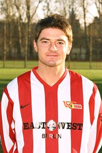 Ein junger Fußballspieler in einem rot-weißen Trikot mit einem Vereinslogo steht vor einem Fußballplatz.