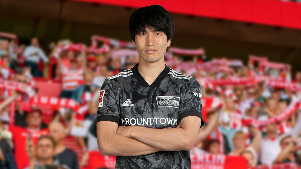 Ein Fußballspieler in schwarzem Trikot steht mit verschränkten Armen vor jubelnden Fans im Stadion.