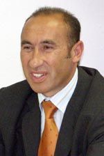 Ein Mann mit kurzen Haaren und Anzug sitzt lächelnd, trägt eine orange Krawatte und hat einen neutralen Hintergrund.