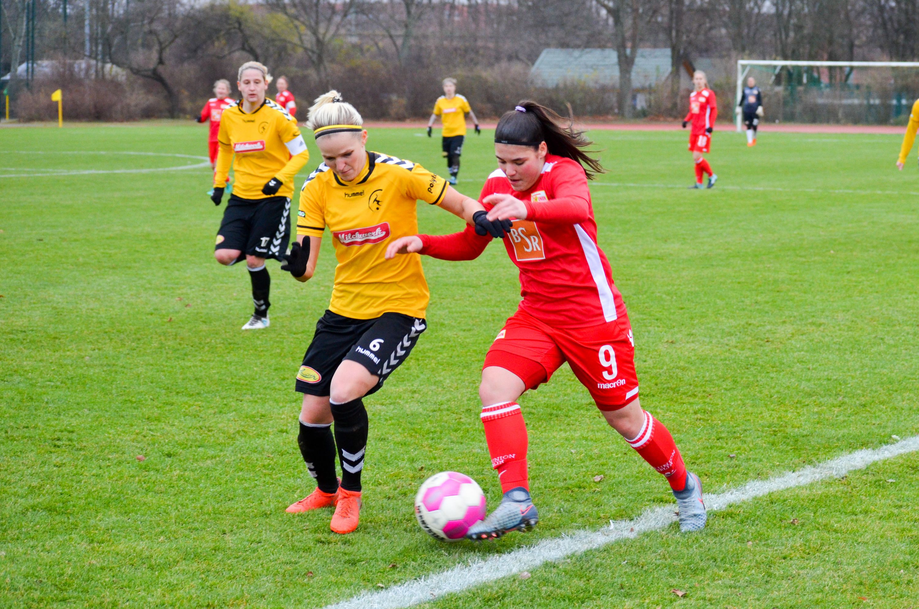 Zwei Fußballspielerinnen kämpfen um den Ball auf einem grünen Spielfeld. Eine trägt ein rotes, die andere ein gelbes Trikot.