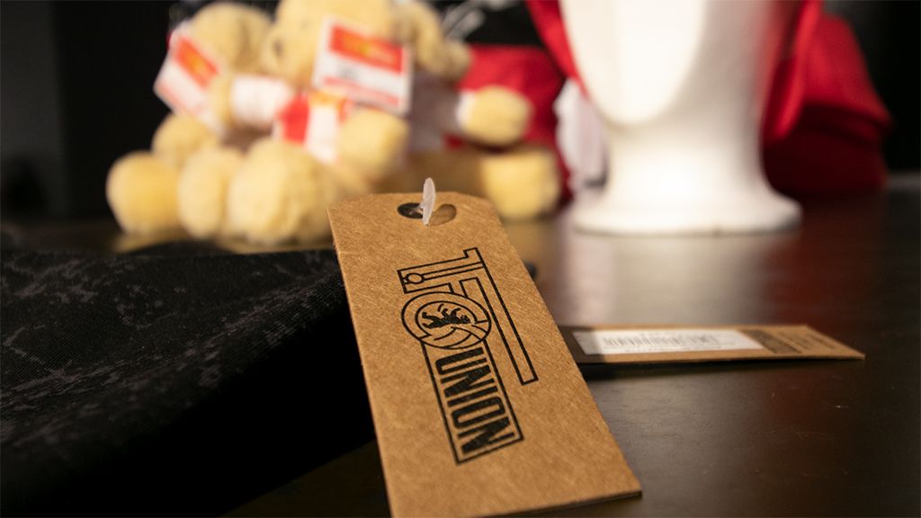 Ein Preisschild mit einem Logo liegt auf einem Tisch, während ein Getränk und zwei Plüschbären im Hintergrund sichtbar sind.
