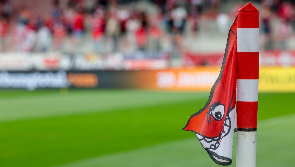 Corner flag with a smiling mascot in front of a stadium with blurred fans in the background.