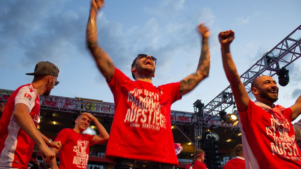 Fans feiern jubelnd in roten T-Shirts, während sie die Hände in die Luft werfen. Ein begeisterter Moment im Stadion.