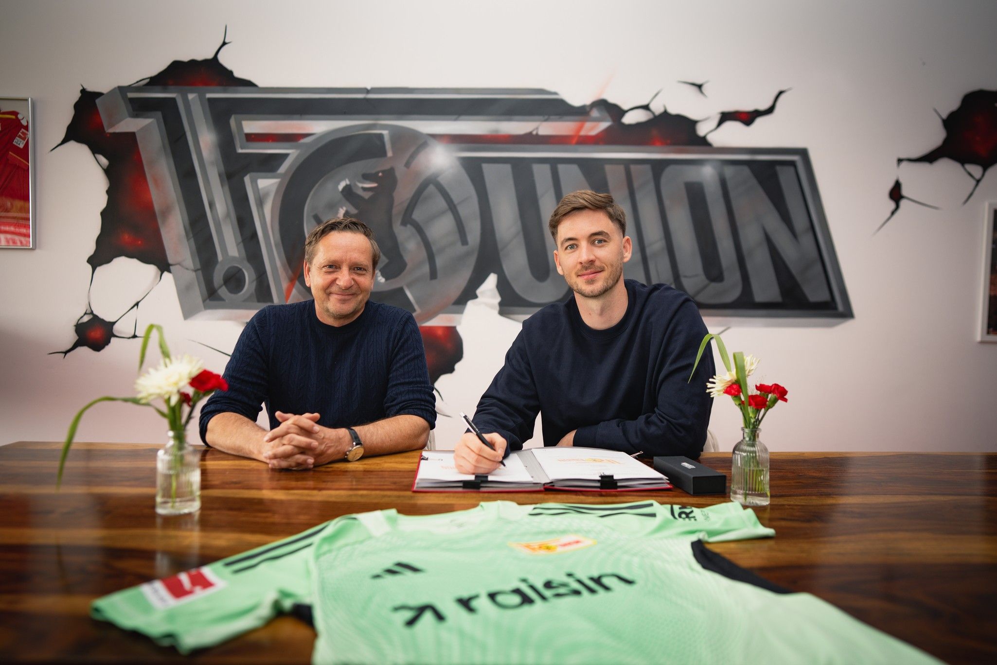 Ein Fußballspieler sitzt am Tisch und unterschreibt einen Vertrag. Im Hintergrund ist das Logo von 1. FC Union zu sehen.
