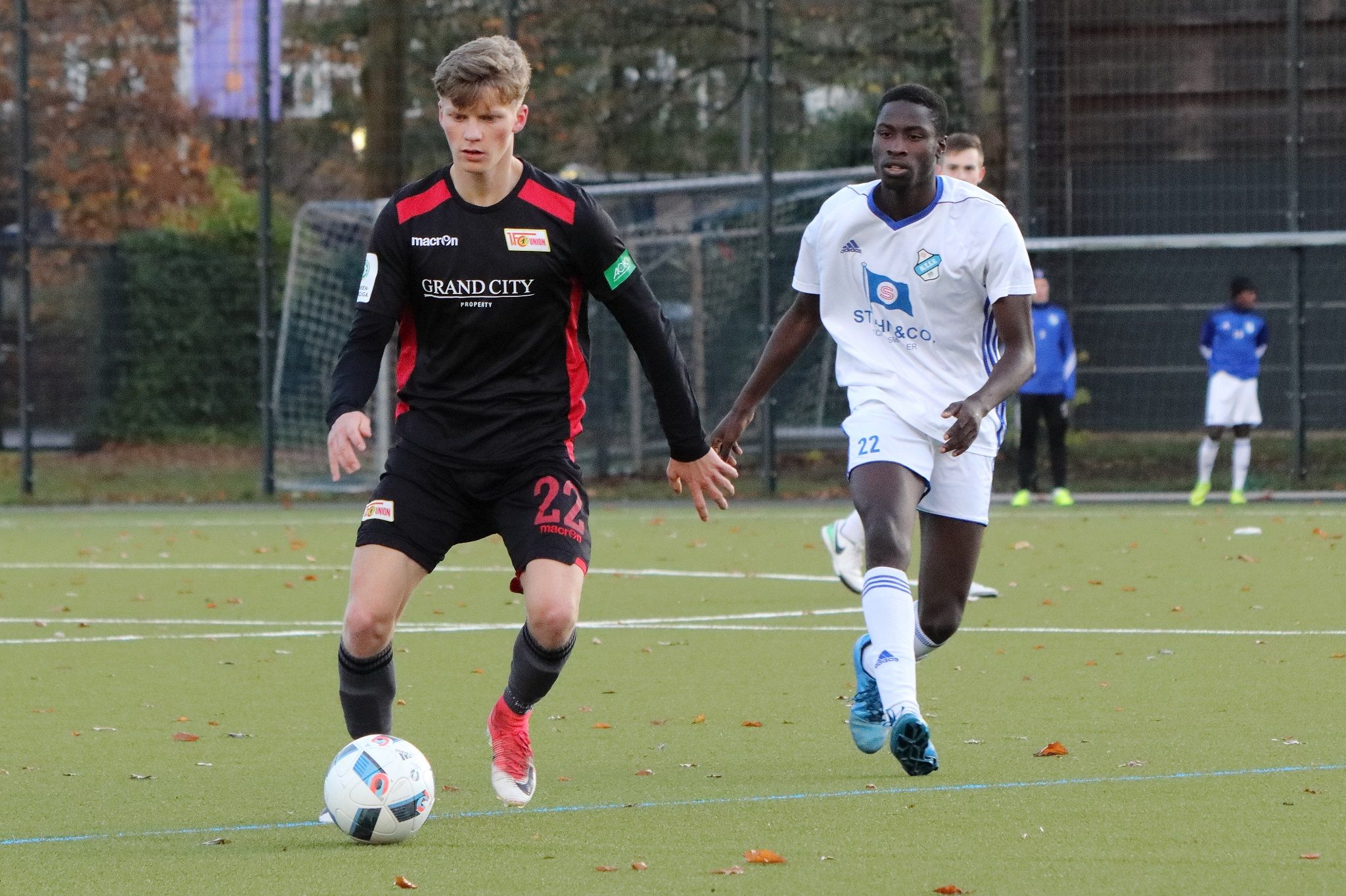 Two soccer players in action: one in a black jersey dribbles the ball while the other in a white jersey follows.