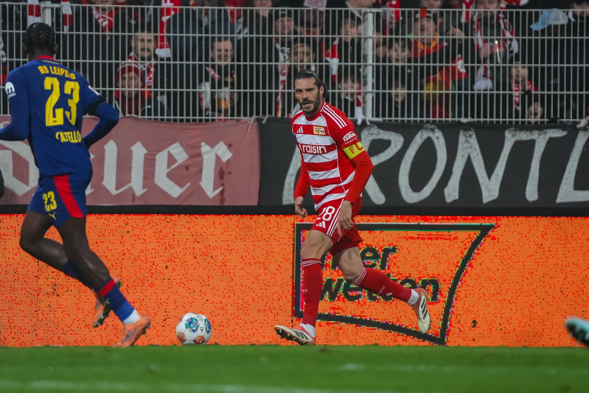 Ein Fußballspieler in roter und weißer Uniform mit der Nummer 18 steht auf dem Spielfeld, im Hintergrund jubelnde Fans.