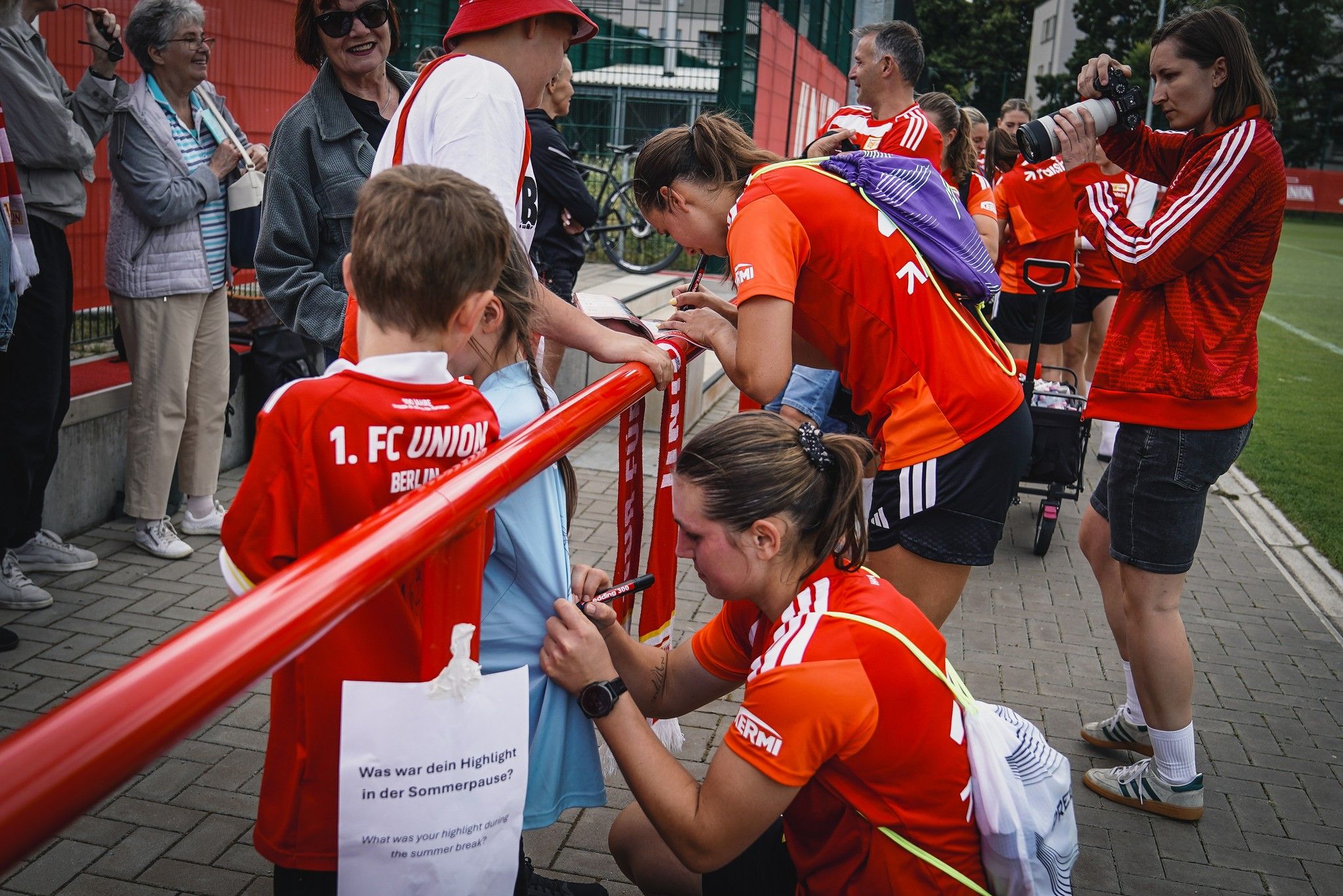 Zwei Frauen in Sportbekleidung autogrammieren auf einem Fußballplatz für Fans in roten Trikots, während andere zuschauen.