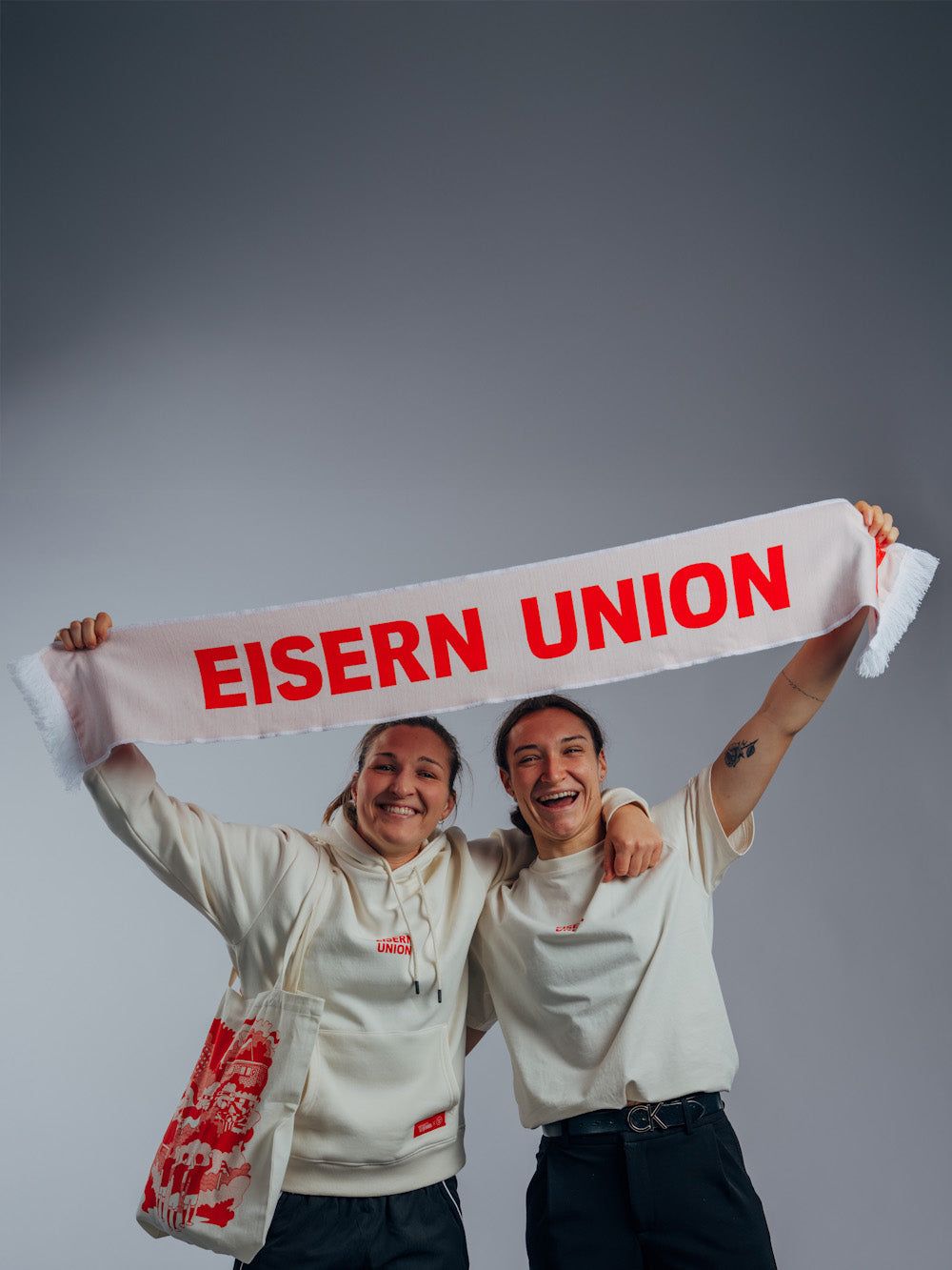 Zwei Personen in hellen Oberteilen halten gemeinsam einen Schal mit der Aufschrift "EISERN UNION" hoch und lächeln fröhlich.
